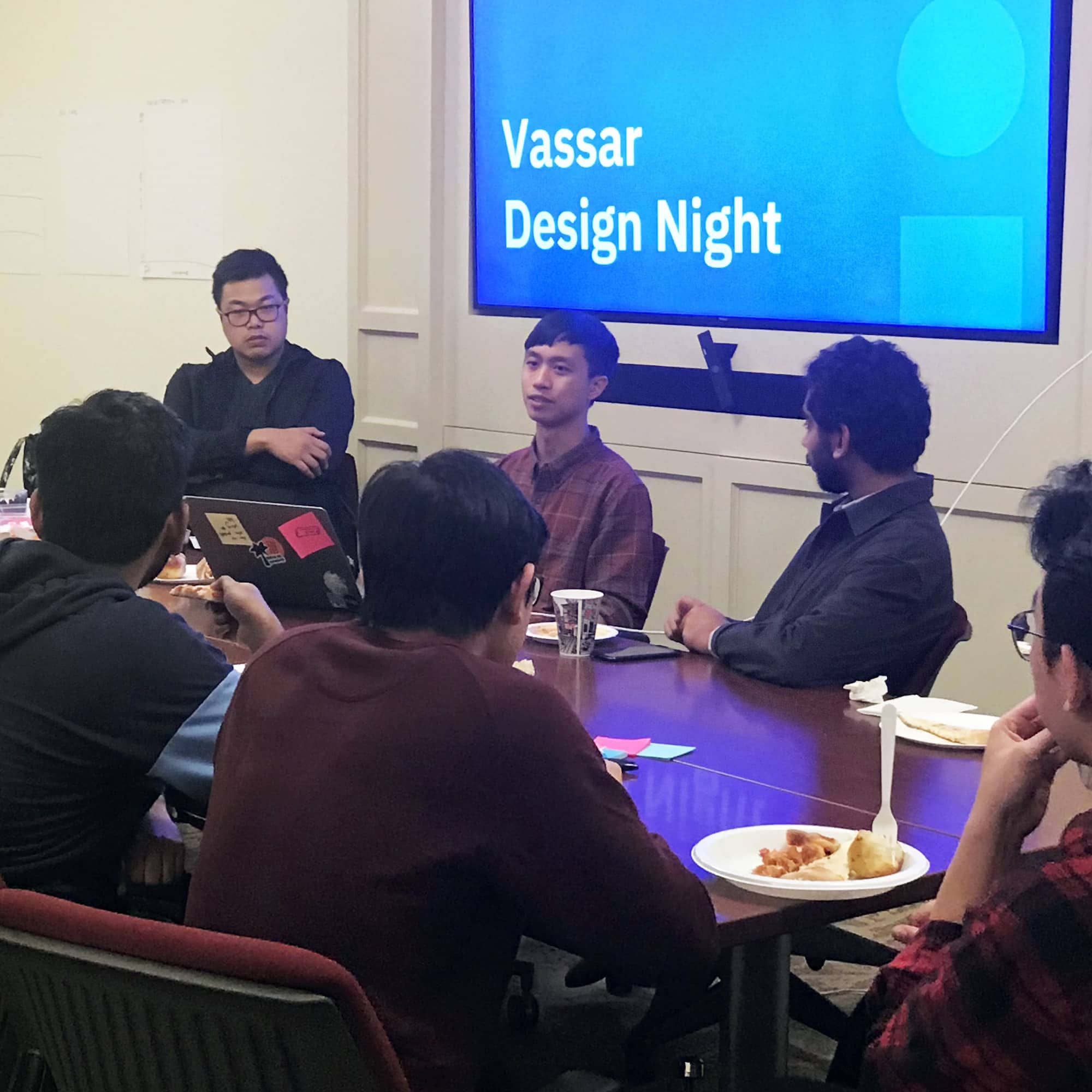 A group of young people are seated around a table, talking, with a projection behind them that reads Vassar Design Night.